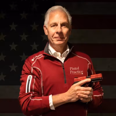 rs=w 1200,h 1200,cg true John Kolis, author of Pistol Practice, wearing a red quarter-zip pullover with the Pistol Practice logo, holding a SIRT training pistol in front of a dark American flag background.