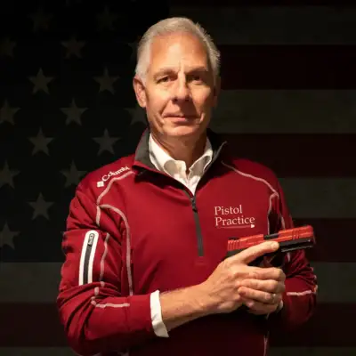 John Kolis, author of Pistol Practice, wearing a red quarter-zip pullover with the Pistol Practice logo, holding a SIRT training pistol in front of a dark American flag background.