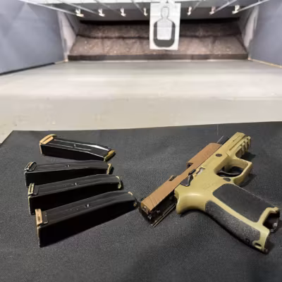 320-downrange Four loaded 9mm magazines and a 9mm semi-automatic with the slide locked to the rear in the foreground. The background features an indoor shooting range with a full-size target downrange, out of focus