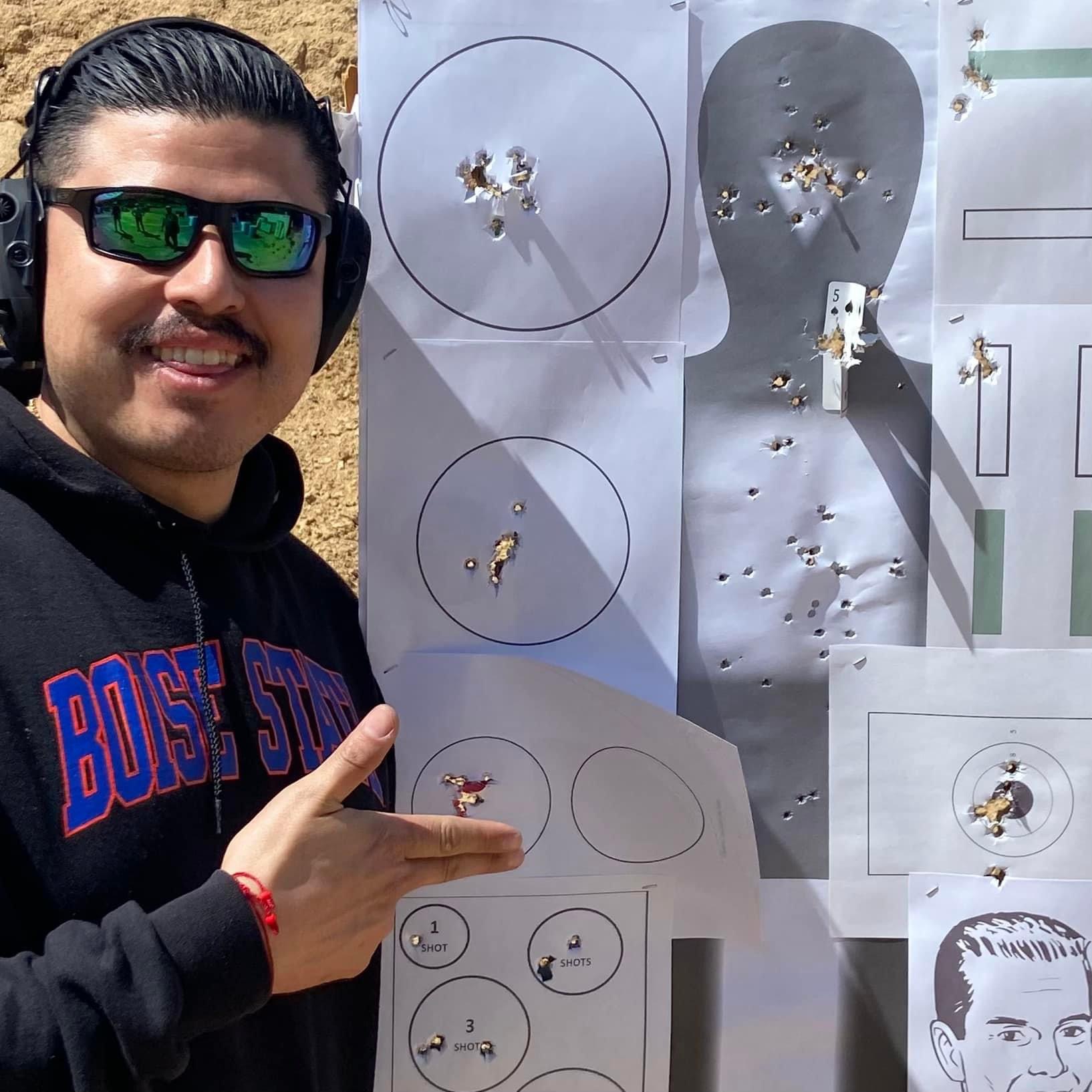 A man facing the camera stands next to his Pistol Practice targets, smiling after completing the Pistol Practice class