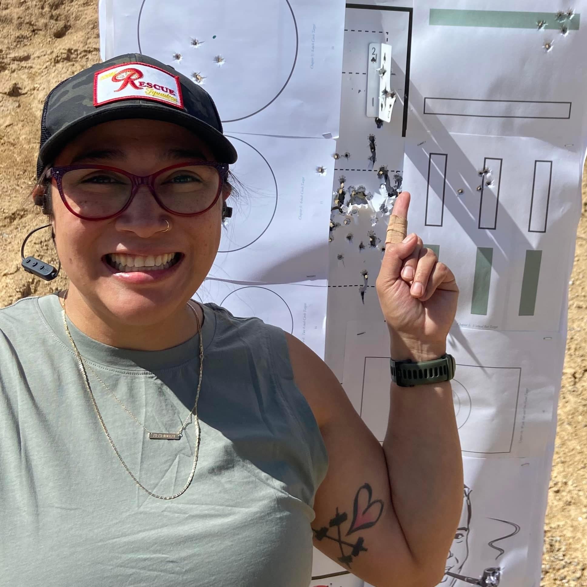 A woman smiling at the camera and pointing to her Pistol Practice targets after just completing the class, showing her successful results