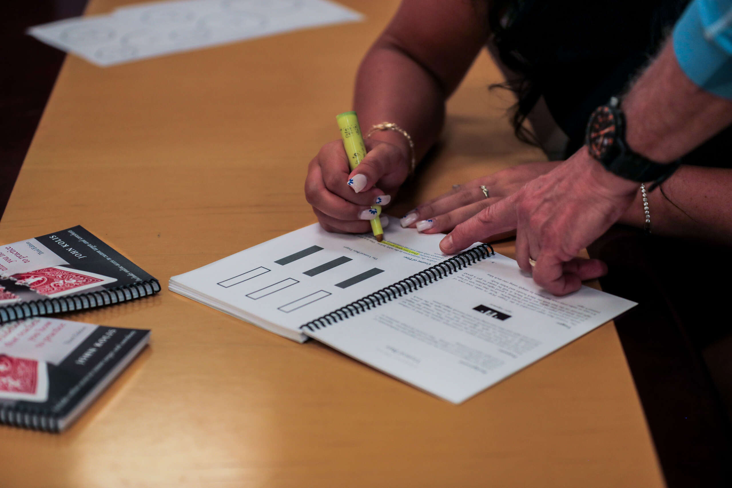 Close-up of a novice highlighting a section in the Pistol Practice book while an instructor points out the text. Only the desk, arms, hands, and books are visible