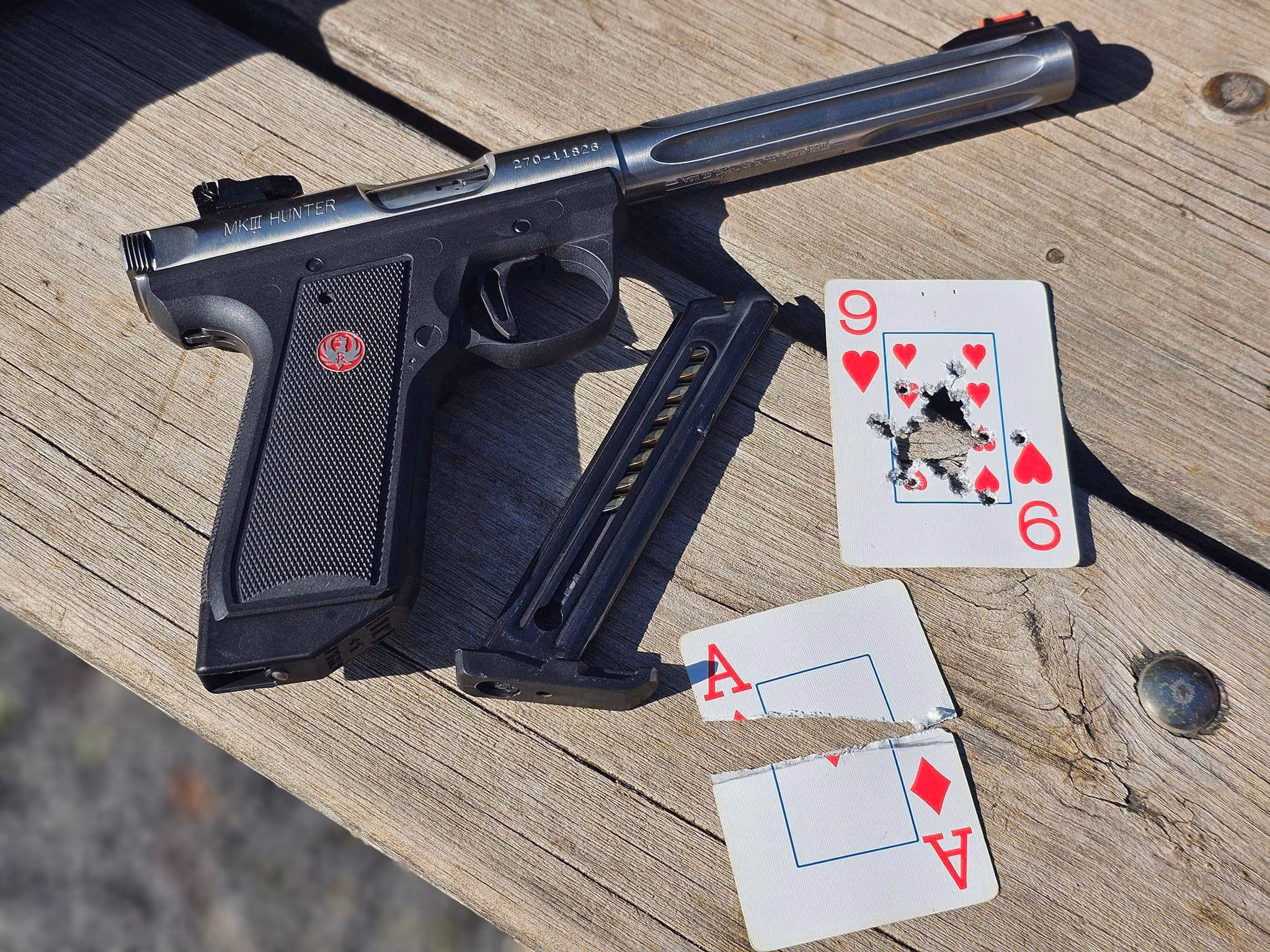 Swanson A pistol lying on a table next to two playing cards that are shot up: an Ace split on edge and a 9 of Hearts with the center shot out
