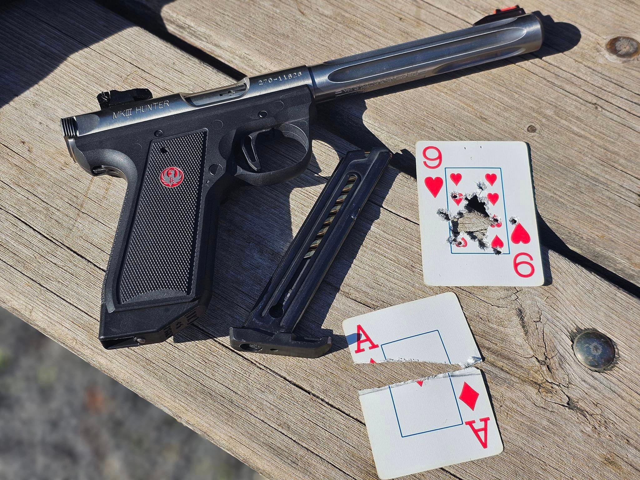 A pistol lying on a table next to two playing cards that are shot up: an Ace split on edge and a 9 of Hearts with the center shot out