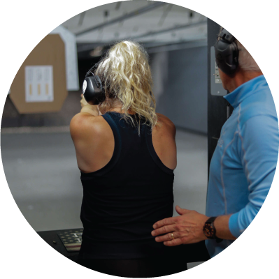 View from behind the firing line of a shooting stall, showing a blonde-haired woman demonstrating good shooting form while a male instructor supervises closely.