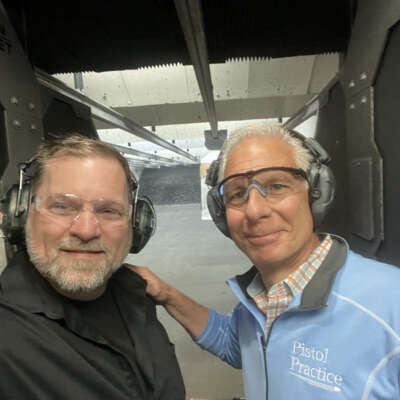 Two men wearing eye and ear protection smiling at the camera with a shooting range stall behind them