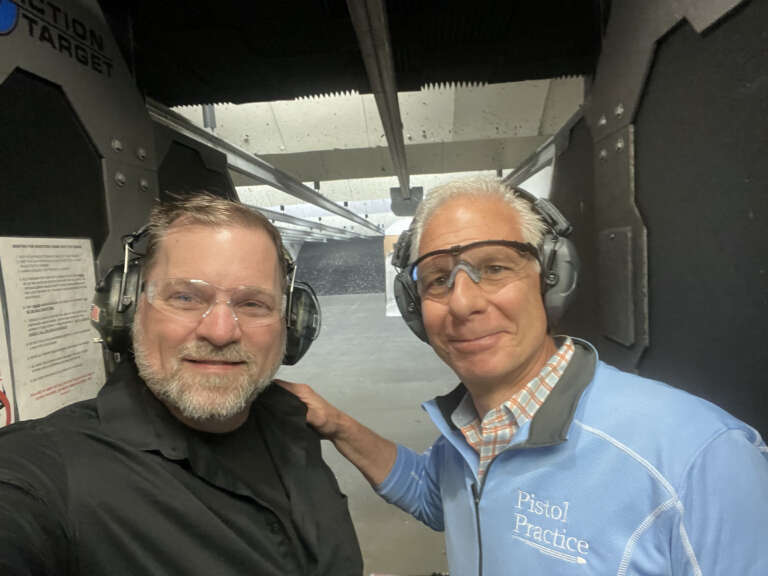 Two men wearing eye and ear protection smiling at the camera with a shooting range stall behind them