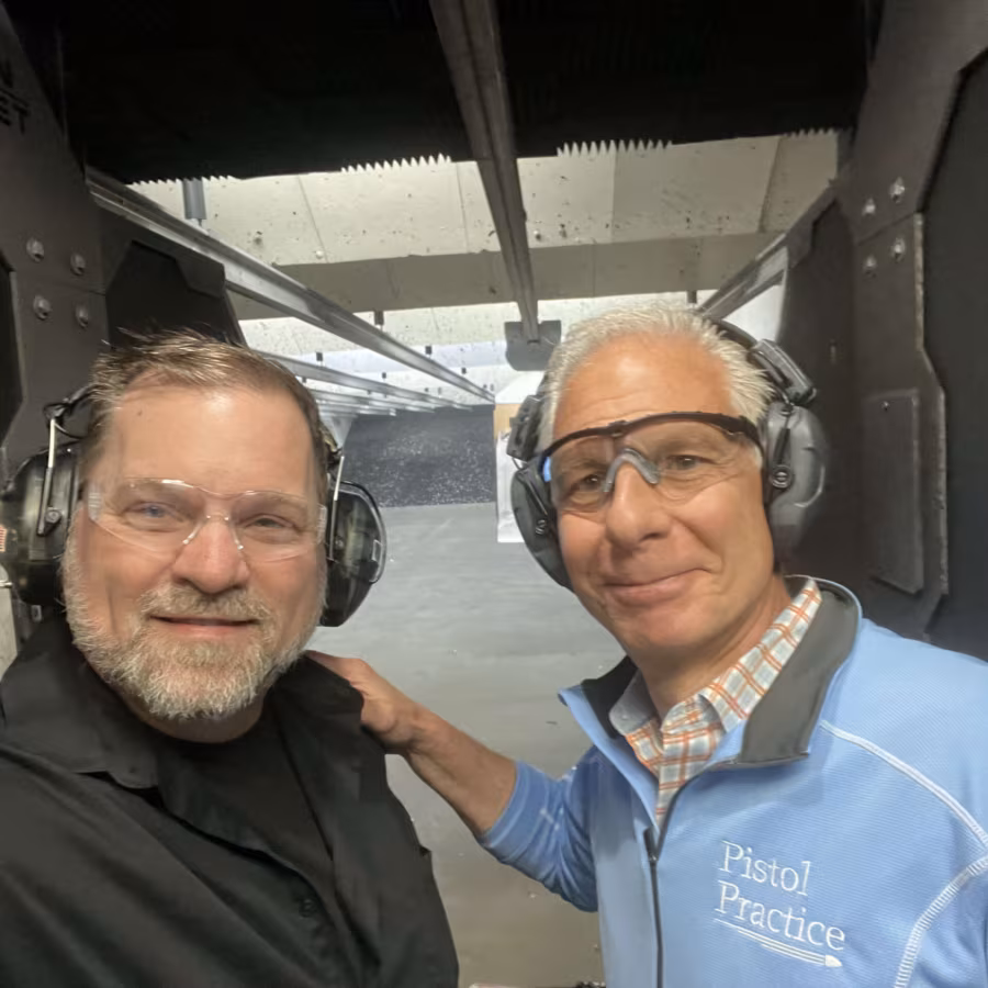 Two men wearing eye and ear protection smiling at the camera with a shooting range stall behind them